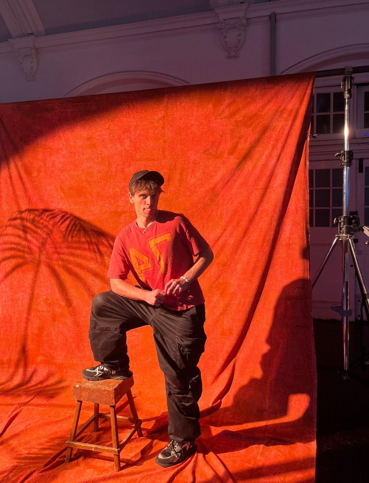 A man poses in a studio setting, wearing a t-shirt, cap, and black pants. The background is a vibrant red, adding a bold contrast to the image.