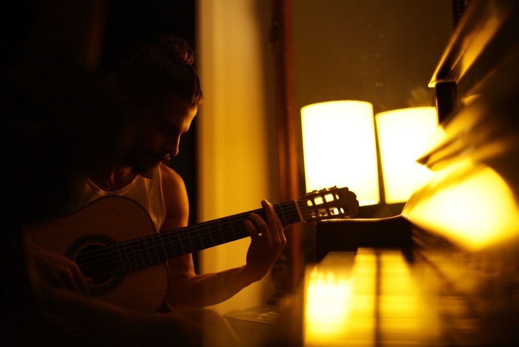 A man plays an acoustic guitar in a dimly lit room. The image is warm-toned, with the guitar and the man's upper body in focus.