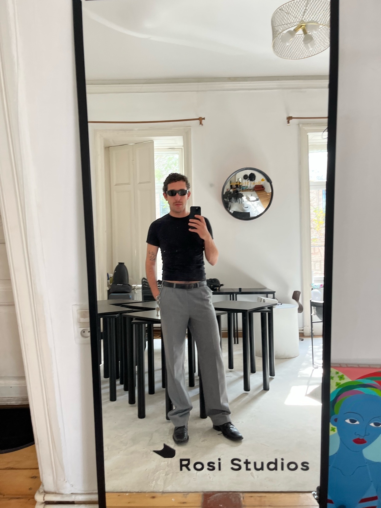 A man takes a mirror selfie, showcasing his outfit. He wears sunglasses, a black t-shirt, and grey pants.
