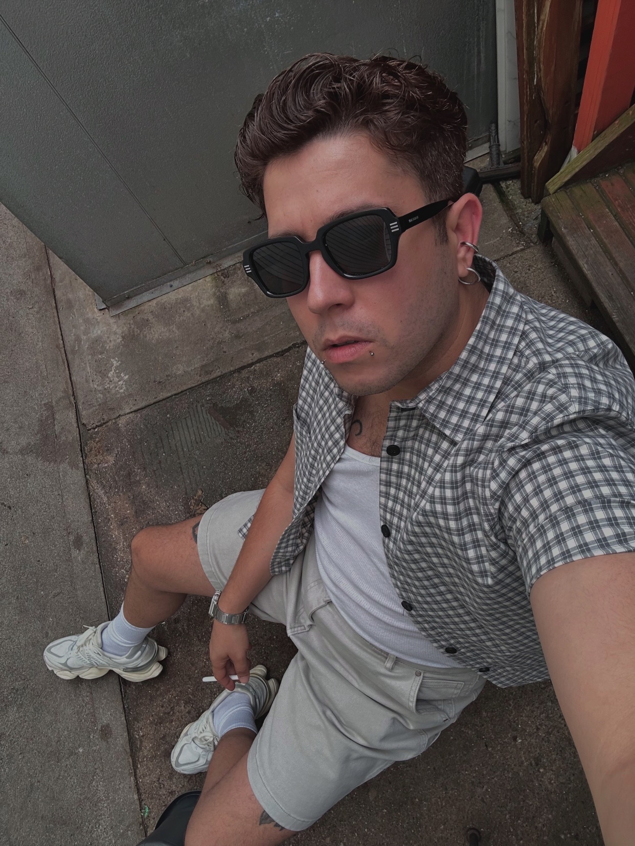 A man poses for a selfie, wearing sunglasses and a stylish outfit. The photo is taken outdoors, with a modern and urban aesthetic.
