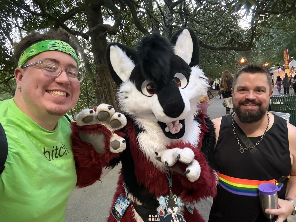 A selfie shows three people posing for a photo. On the left is a man wearing glasses and a green t-shirt. In the middle is a person in a furry costume of a black and white wolf. On the right is a man wearing a black tank top with a rainbow pride flag