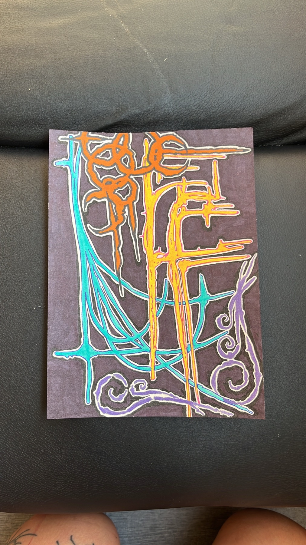 A colorful abstract drawing with swirling lines and shapes in orange, yellow, teal, and purple on a dark purple background. The artwork appears to be placed on a dark surface, possibly a couch.