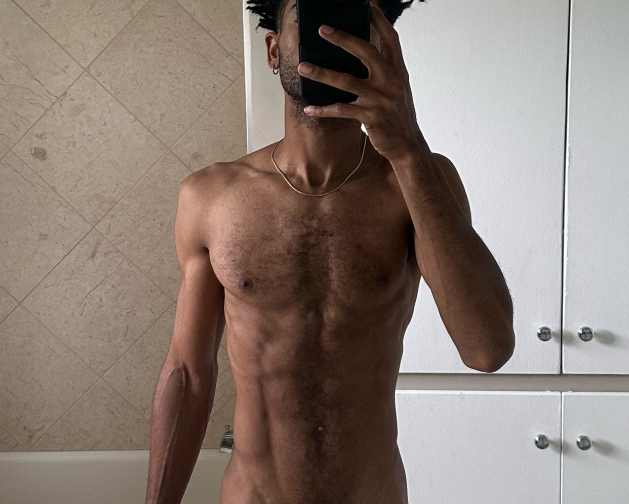 A shirtless man takes a mirror selfie in a bathroom. He has a muscular physique and is wearing a gold necklace.