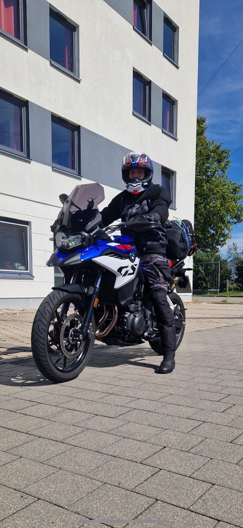 A person wearing a helmet and riding gear sits on a blue and white BMW GS motorcycle parked on a paved surface in front of a modern building.