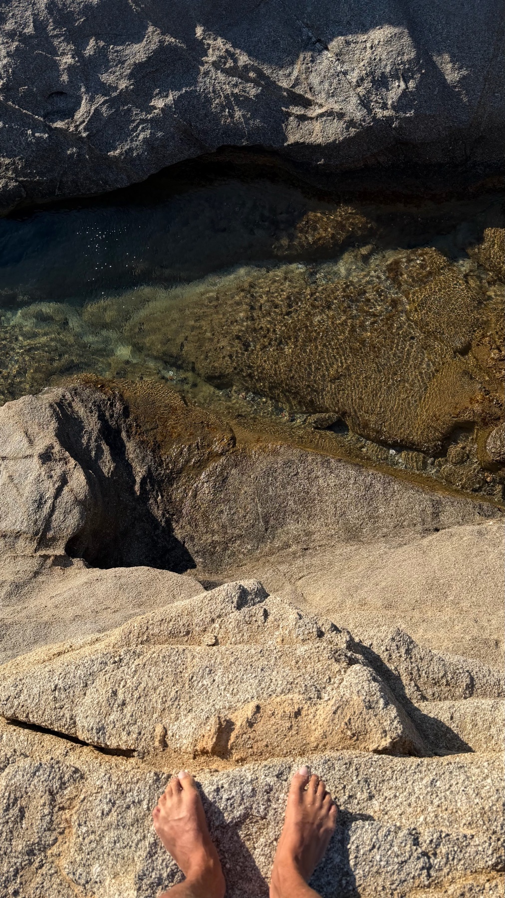 A close-up shot of a person's bare feet standing on a rocky shore with clear water visible in the background.