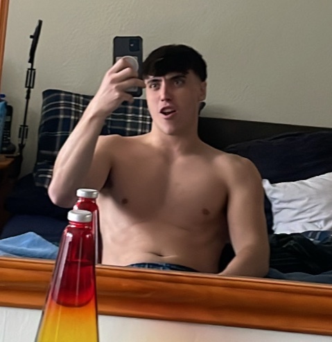 A young man with dark hair takes a selfie in a bedroom, looking surprised. He is shirtless and wearing blue jeans.