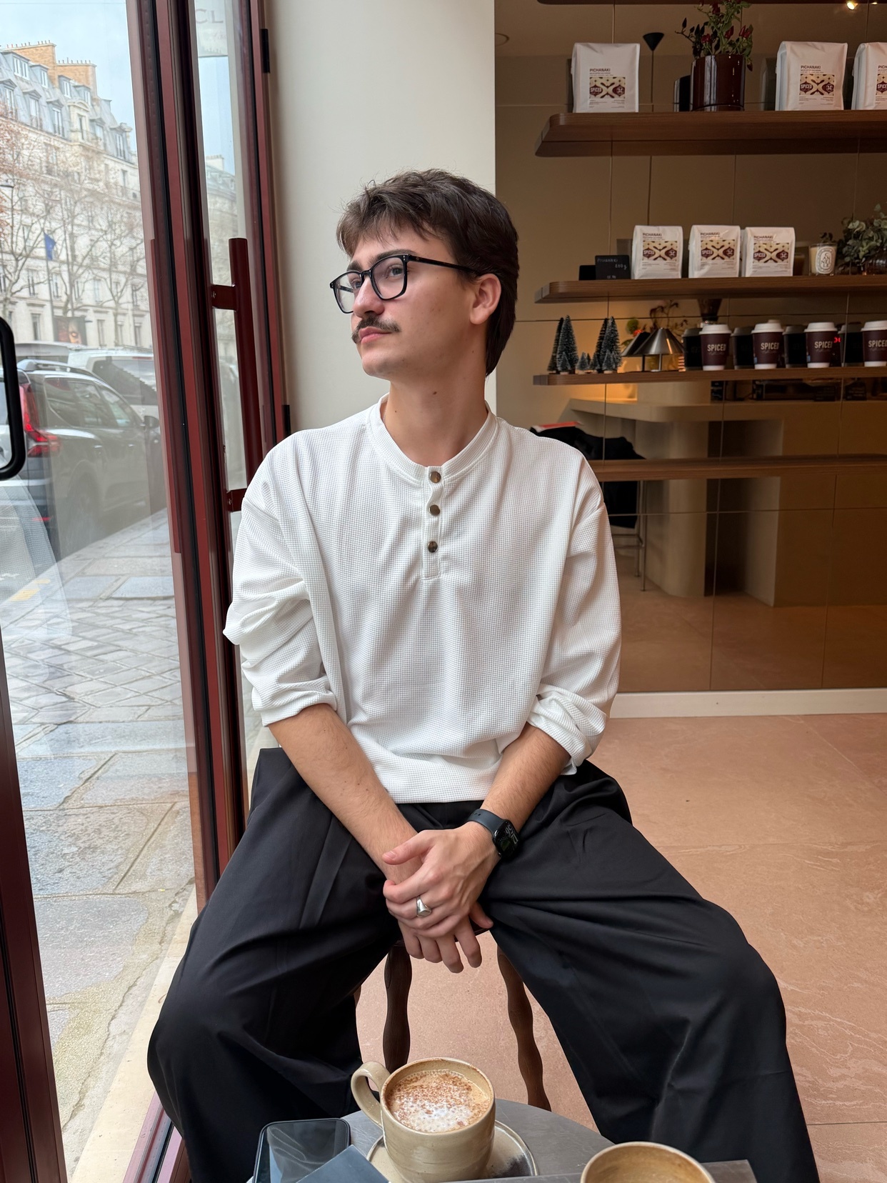 A man sits in a cafe, looking out the window. He wears a white shirt and black pants.