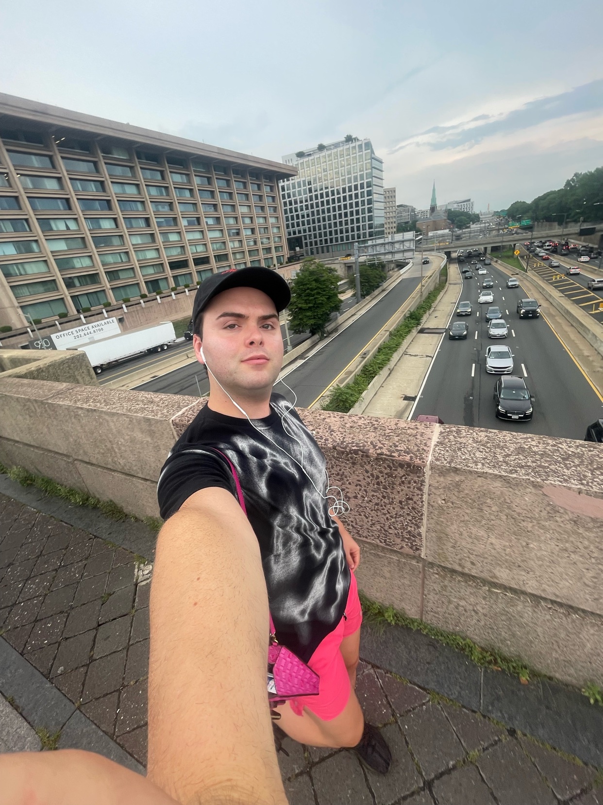 A man takes a selfie on a bridge overlooking a busy highway in a city. He is wearing a black t-shirt, pink shorts, and a black baseball cap.