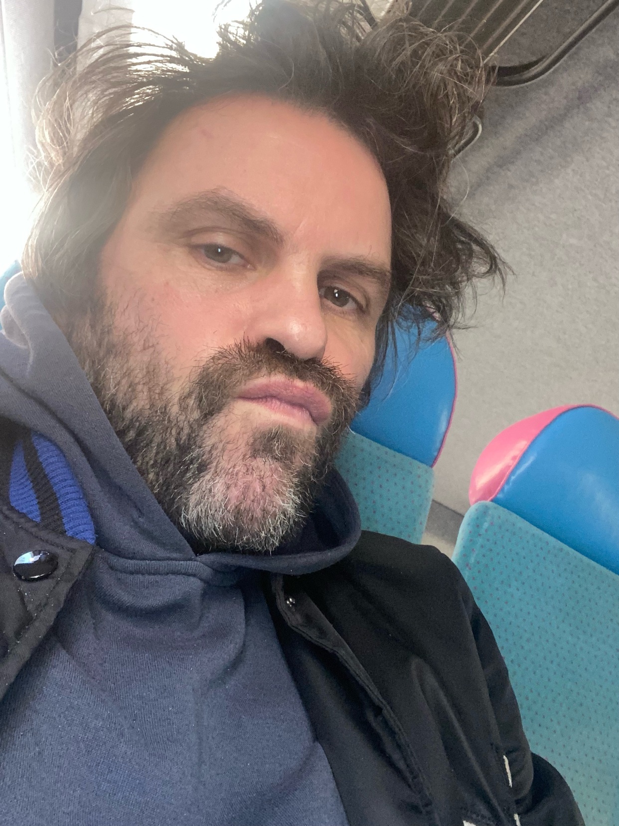 A man with a beard and stubble takes a selfie on a train. His dark hair is messy and he is wearing a hoodie and jacket.