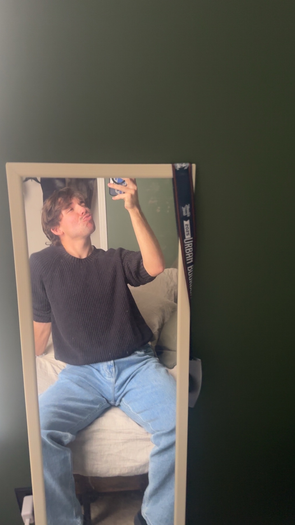 A man takes a mirror selfie, showcasing his outfit. He's wearing a black shirt and blue jeans.