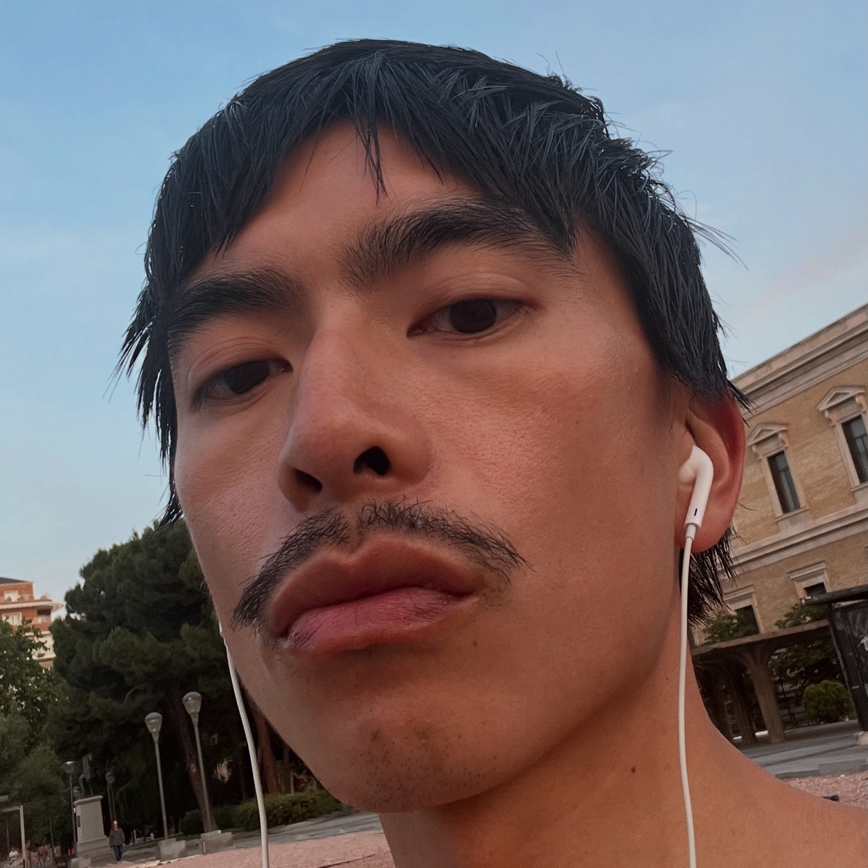 A selfie of a man with dark hair and a mustache. He is shirtless and wearing earphones.