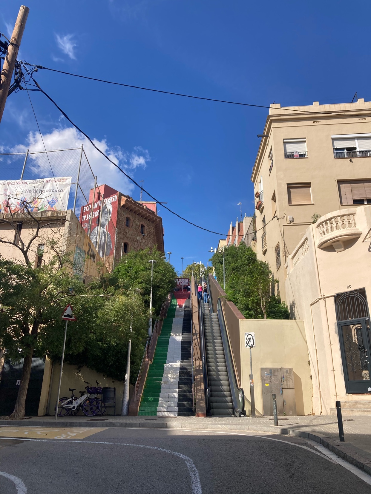 An outdoor escalator ascends a street decorated with watermelon-themed street art. The steps are painted green, white, and black, resembling the fruit, under a bright blue sky.