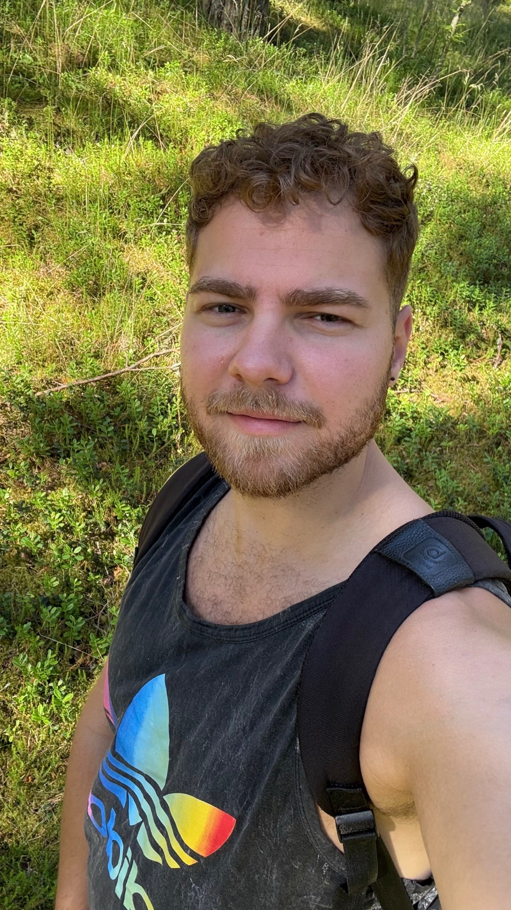 A man with curly brown hair and stubble smiles at the camera while wearing a dark tank top with an Adidas logo. He is outdoors with green foliage in the background.
