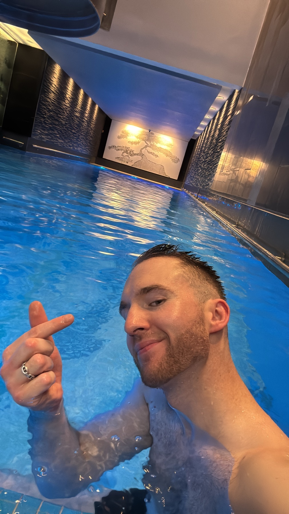 A man with short hair and stubble smiles at the camera while making a finger heart gesture in a swimming pool. The water is blue and there are lights reflecting off the surface.