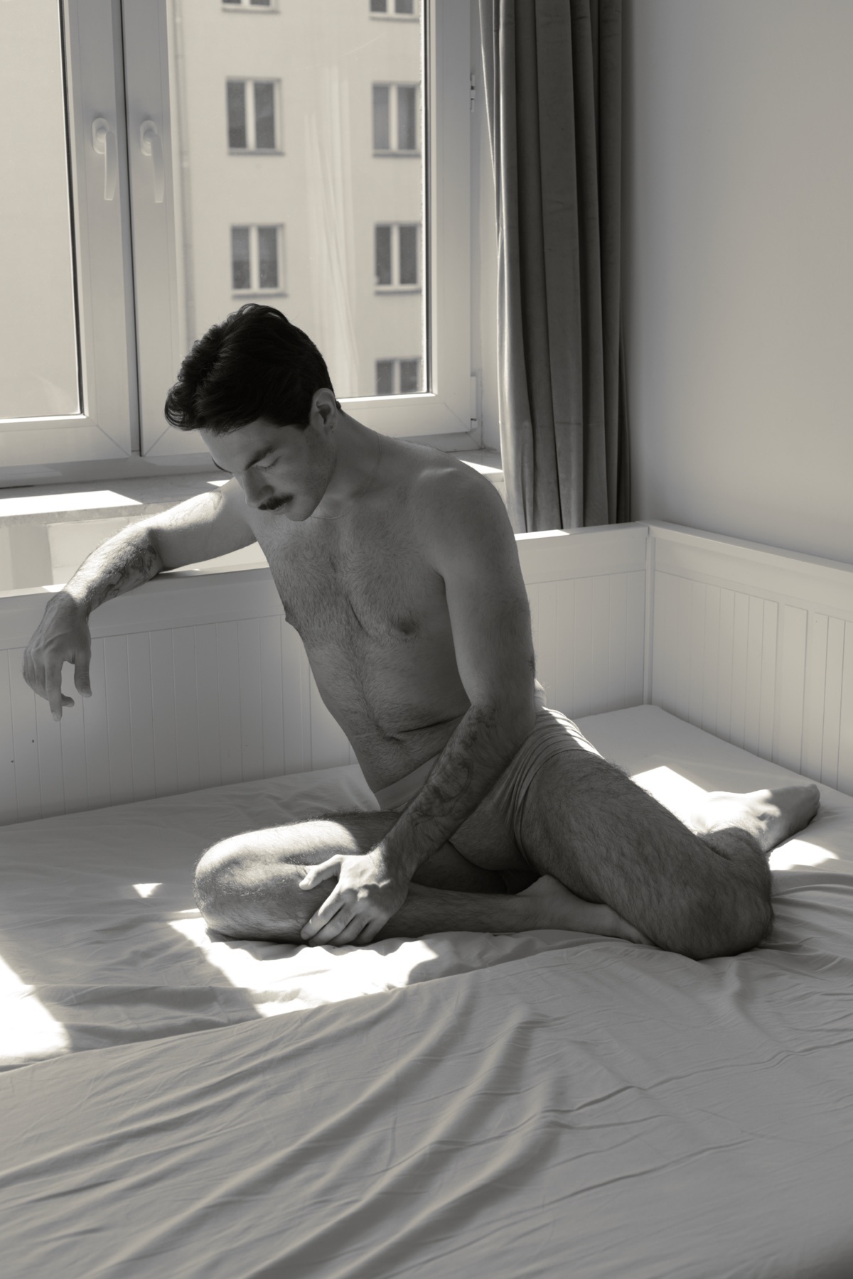 A shirtless man with a mustache and tattoos sits on a bed in a room with natural light streaming in from a window. He is wearing underwear and appears to be in a contemplative pose.