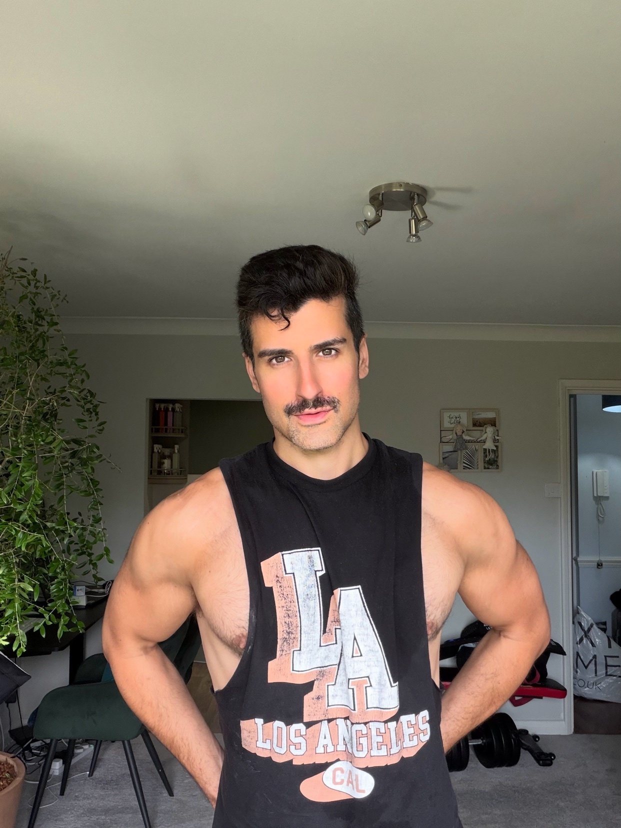 A man with a mustache poses indoors wearing a black tank top with 'LA' and 'Los Angeles' printed on it. He has muscular arms and is looking directly at the camera.