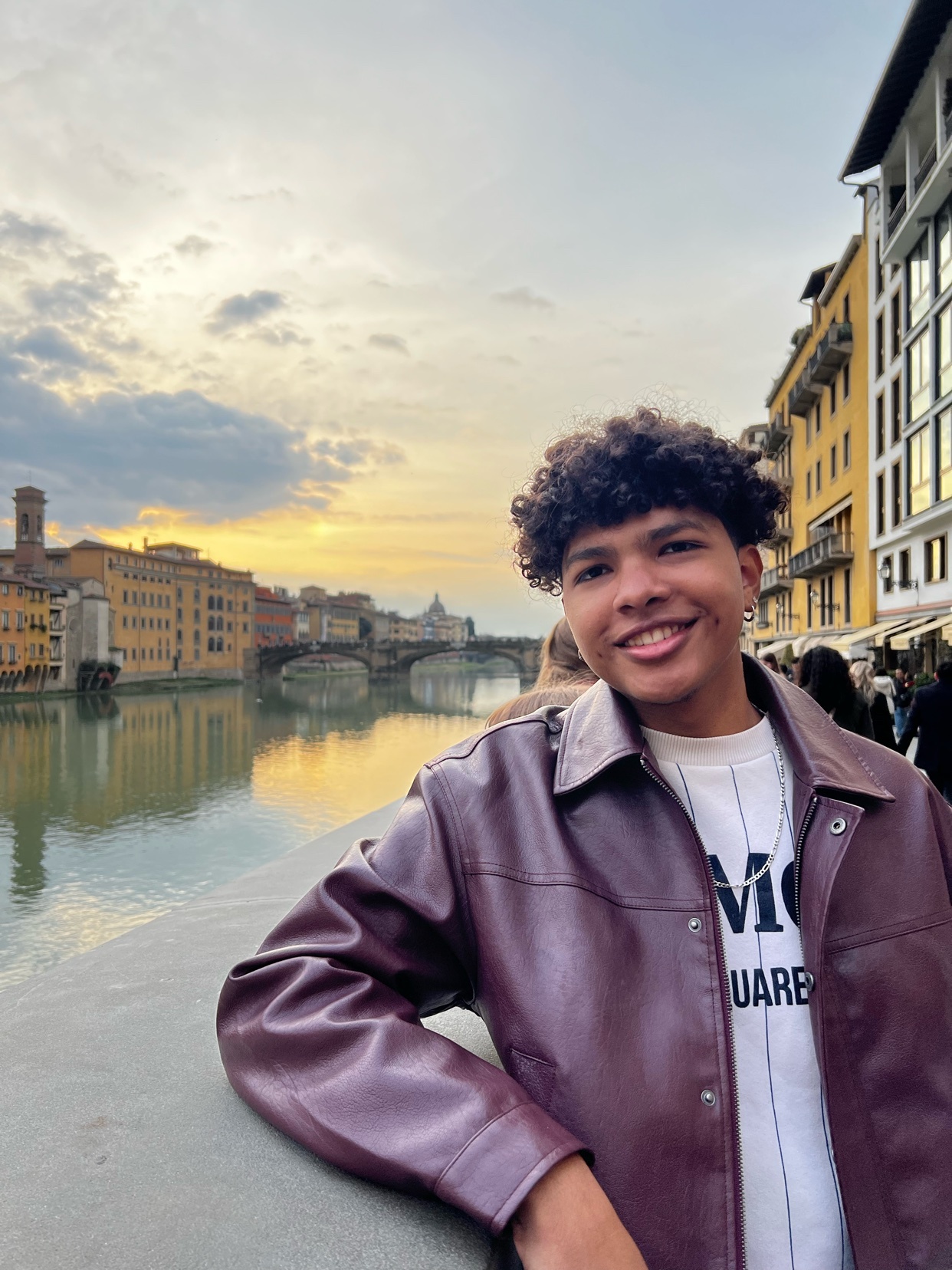 A young man with curly hair smiles at the camera, wearing a leather jacket and a necklace. He is standing on a bridge with a river and historic buildings in the background.