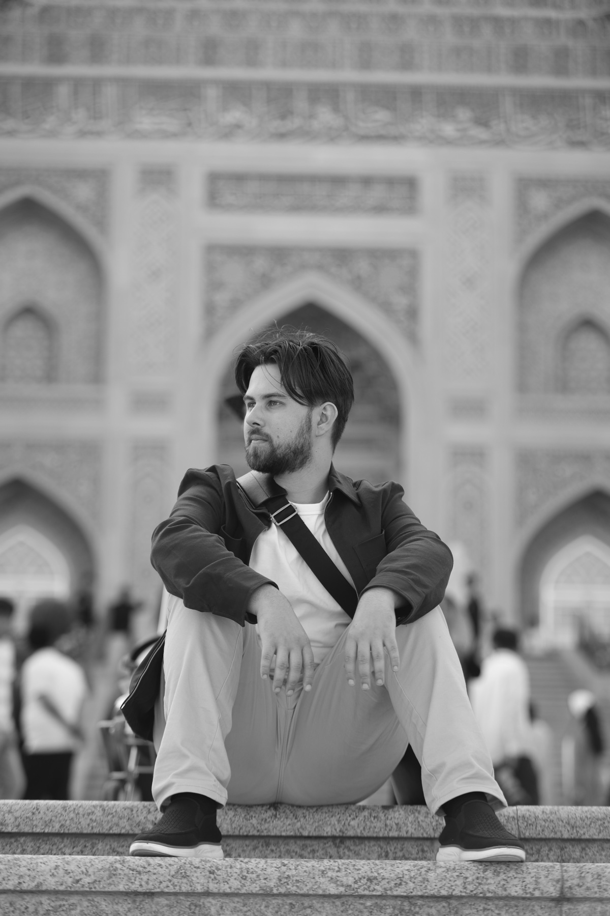 A black and white photo shows a man with a beard sitting on steps in front of an ornate building. He is wearing a jacket and pants and looking off to the side.