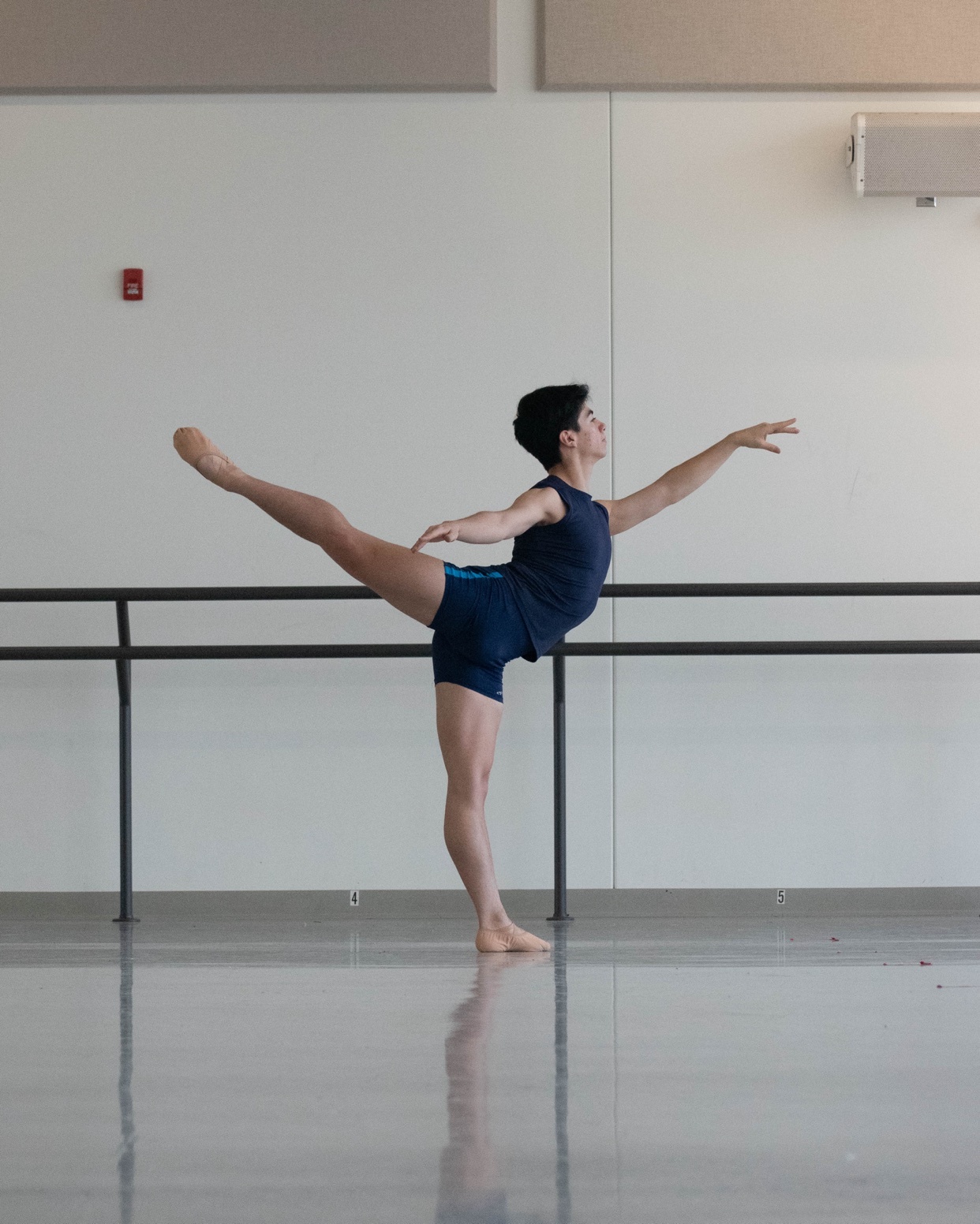 A man in athletic wear performs a ballet pose in a dance studio. The image captures the dancer's form and the studio's minimalist aesthetic.