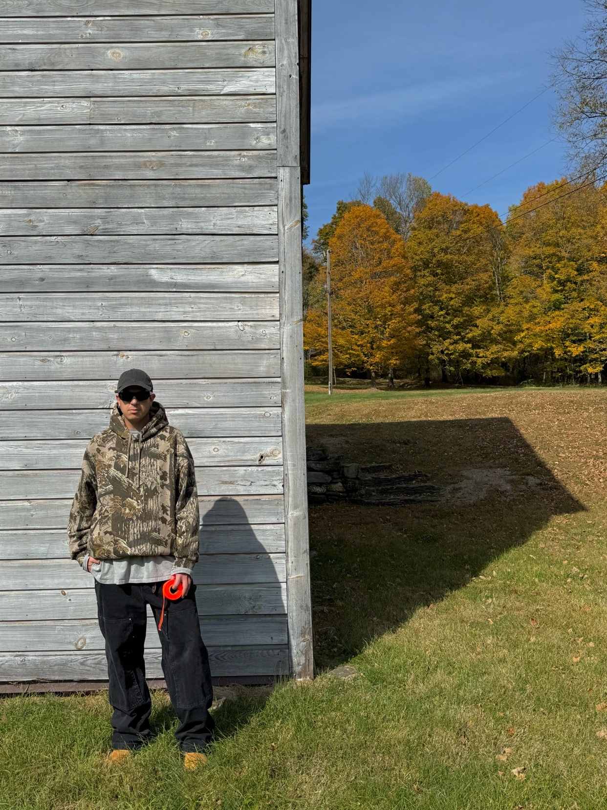 A man stands outside in front of a wooden building. He is wearing a camouflage hoodie and sunglasses.