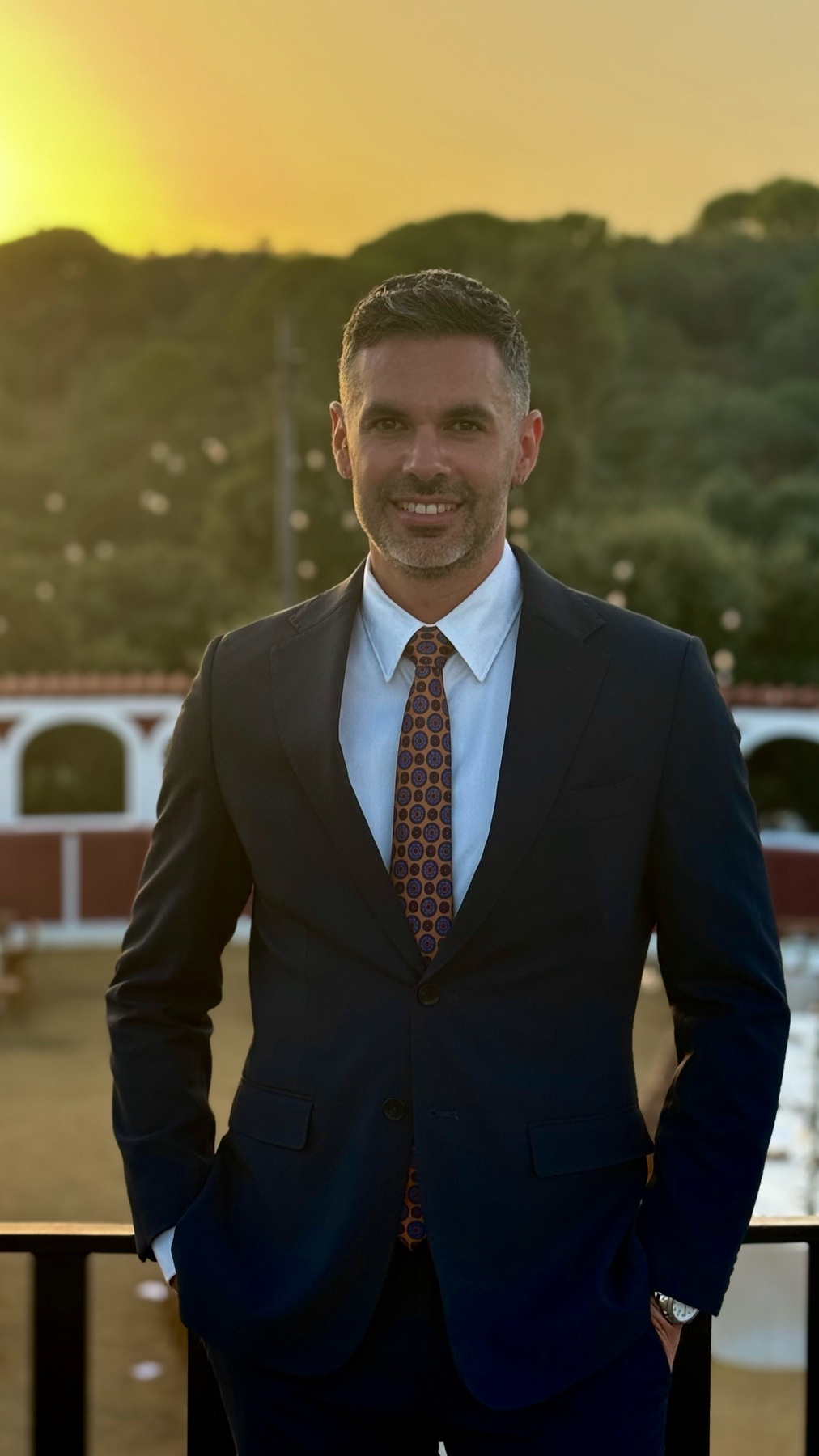 A man in a dark suit, white shirt, and patterned tie smiles at the camera with his hands in his pockets. The background features a warm sunset over a hilly landscape.