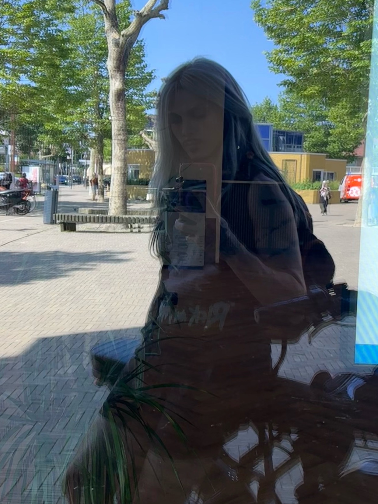 A woman with long, two-toned hair takes a selfie in a reflective window on a sunny day. She has tattoos visible on her arms.