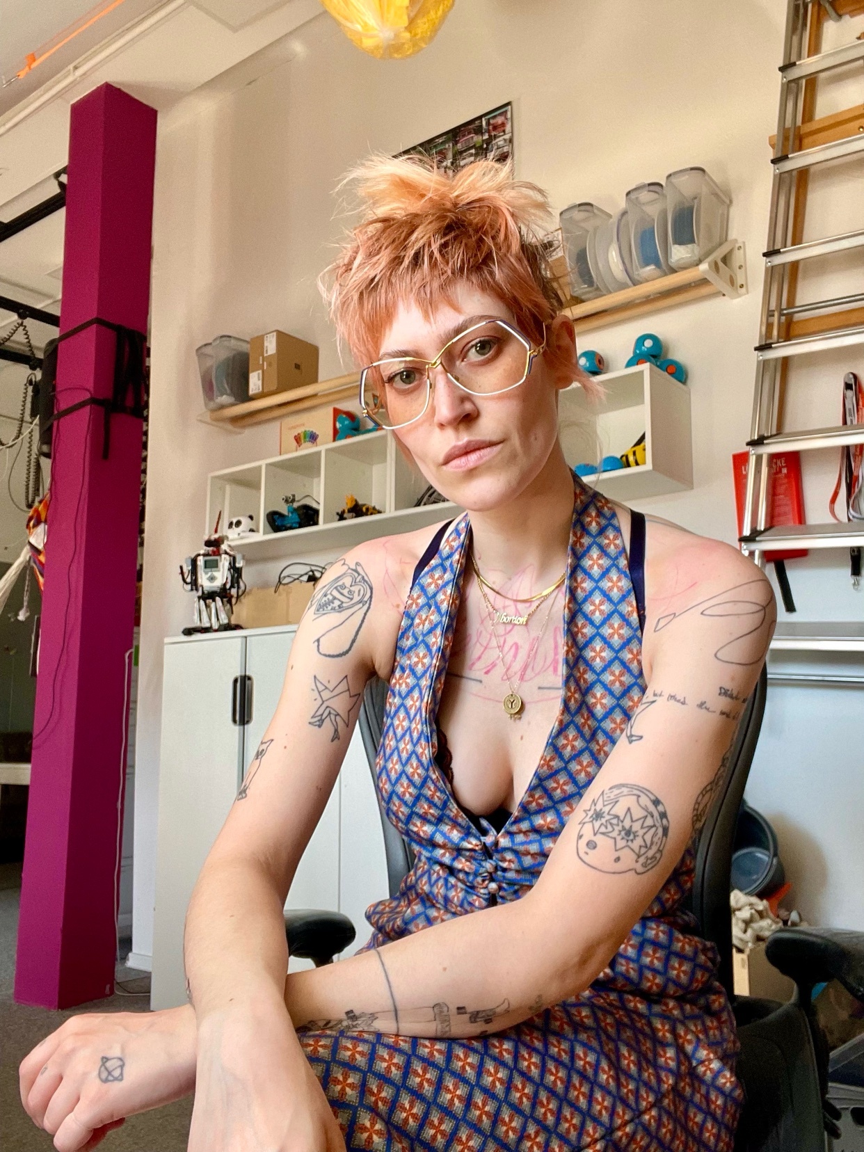 A woman with short, spiky pink hair and glasses poses in a blue patterned dress. She has tattoos on her arms and a serious expression.