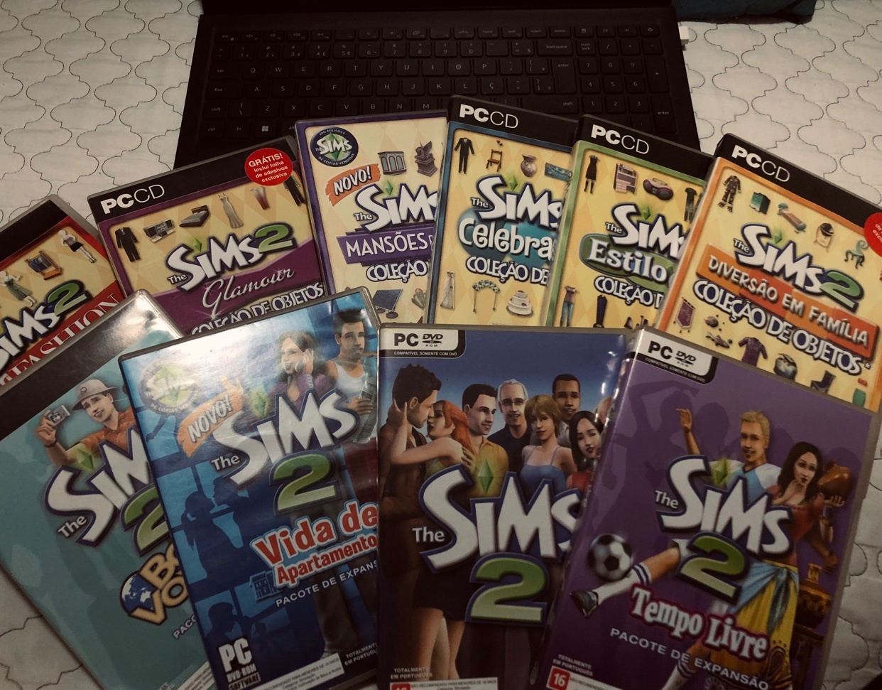 A collection of Sims 2 PC game expansion packs are displayed on a laptop keyboard. The game cases feature colorful artwork and titles like 'Glamour,' 'Celebration,' and 'Family Fun.'