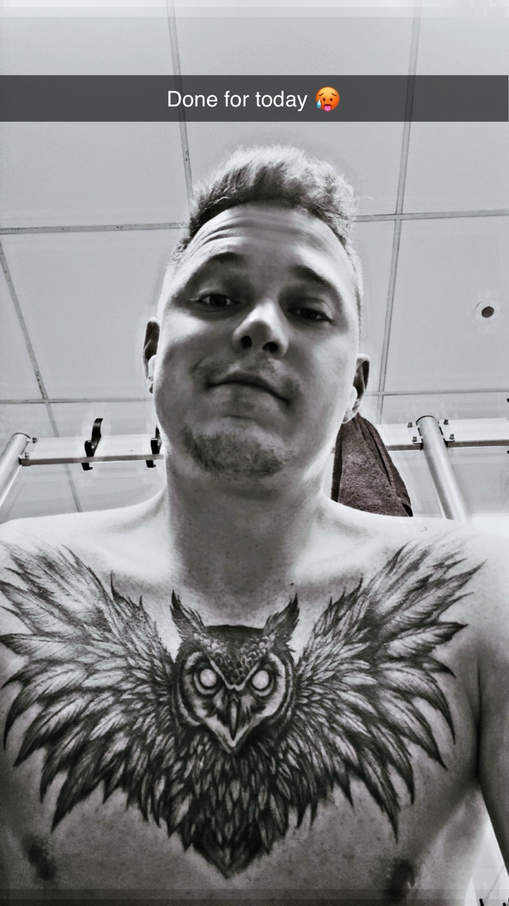 A black and white selfie of a man with a large owl tattoo on his chest. He is looking at the camera with a slight smirk.