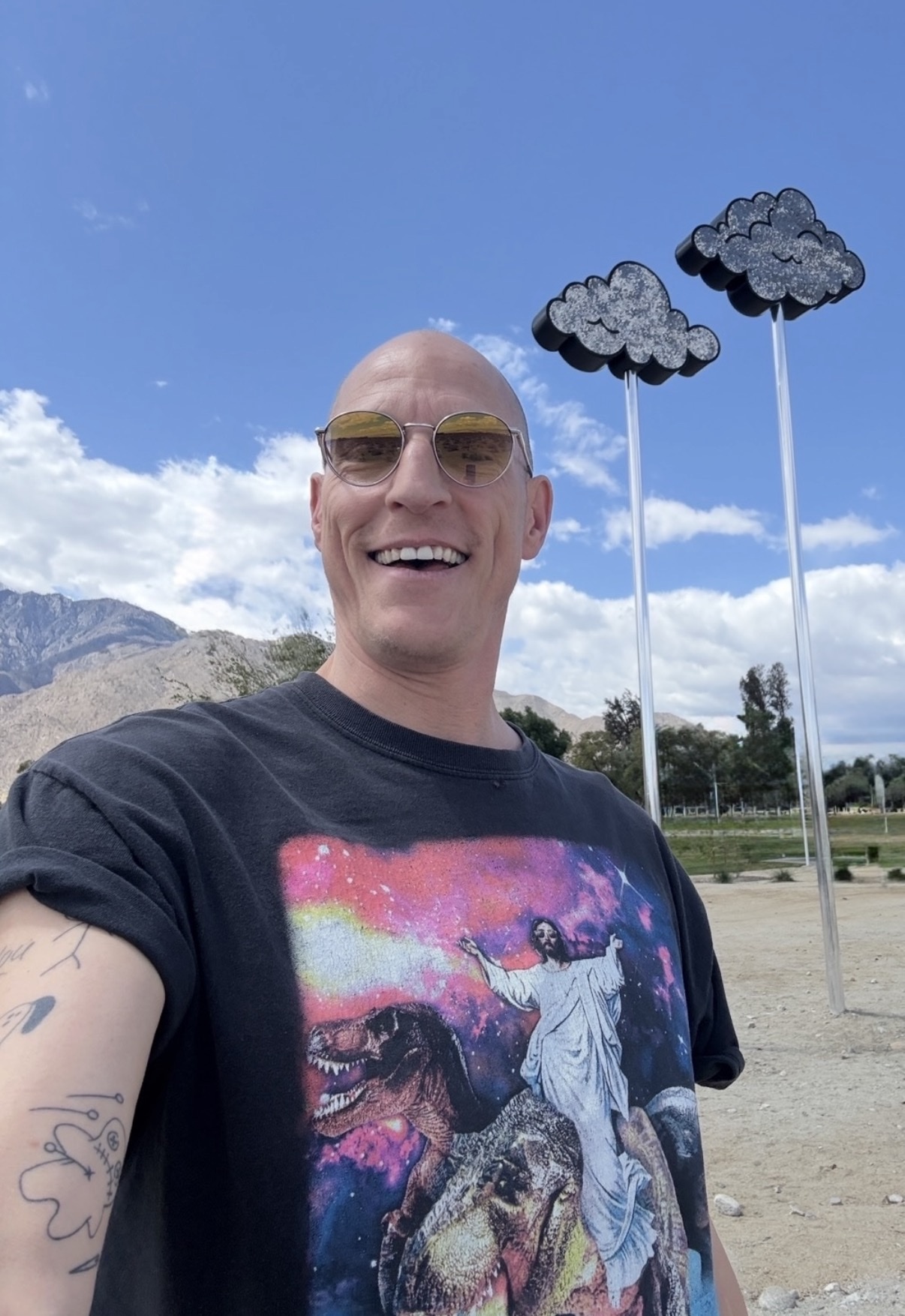 A bald man with sunglasses and a graphic t-shirt smiles for a selfie outdoors. Behind him, two cloud sculptures stand tall against a bright blue sky with mountains in the distance.