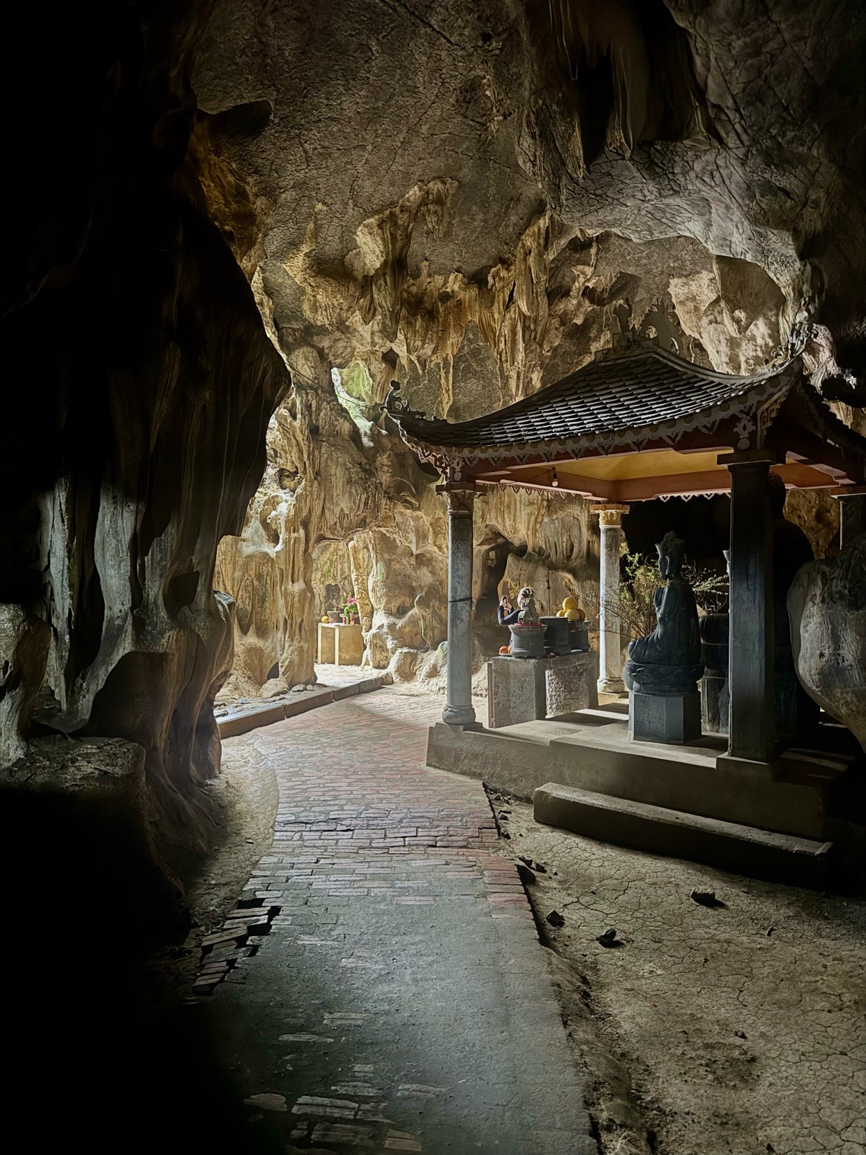 A dimly lit cave interior features a brick pathway leading to a small, ornate pagoda housing a Buddha statue. The rough, textured walls of the cave contrast with the man-made structure, creating a serene and ancient atmosphere.