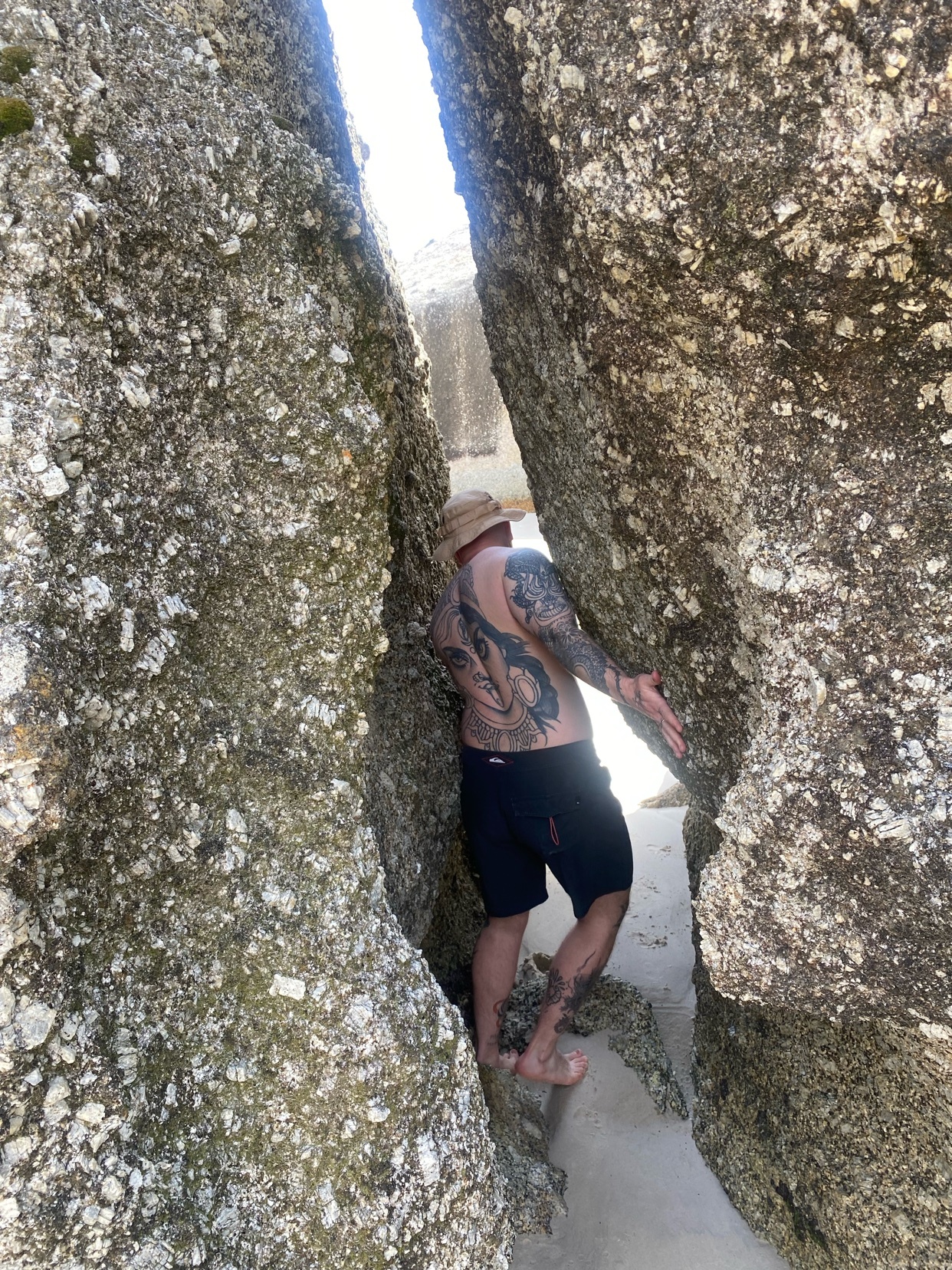A man with a straw hat and extensive tattoos on his back and legs is squeezing himself between two large rocks on a beach.