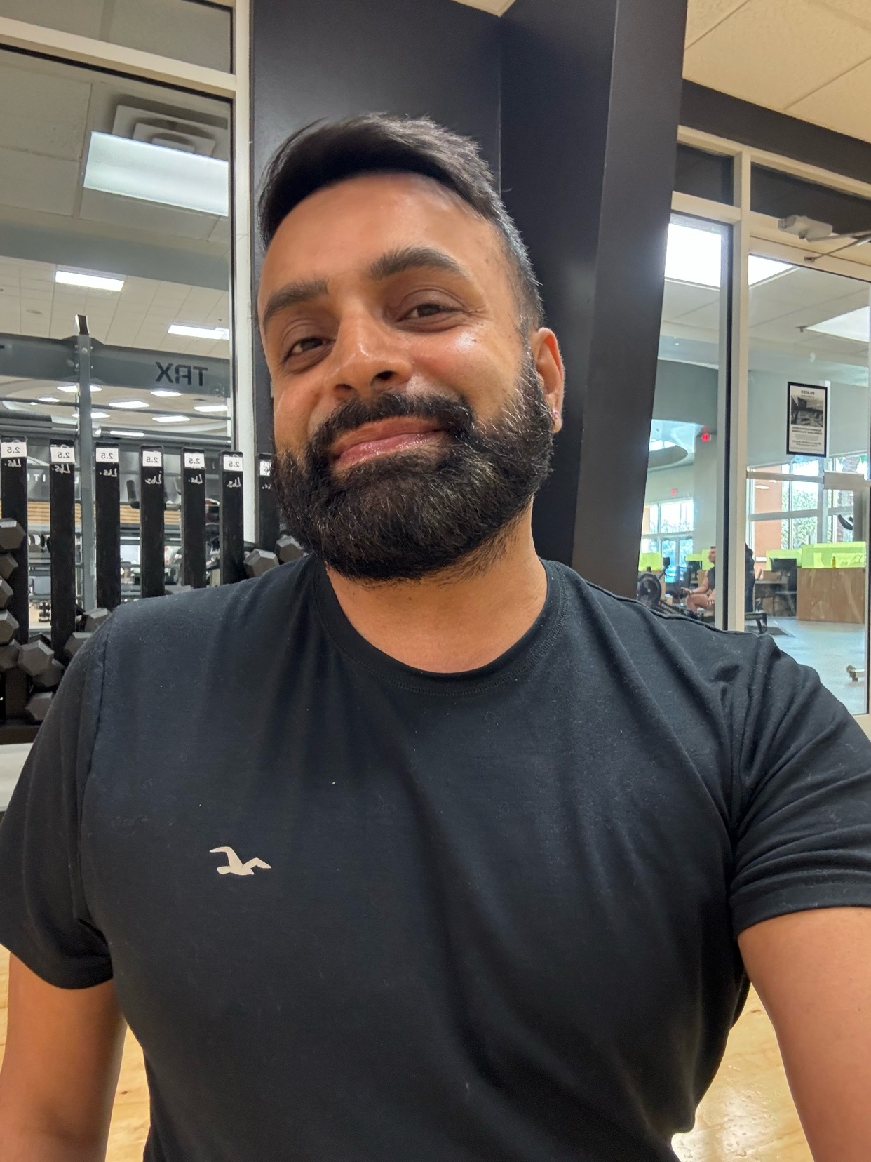 A man smiles at the camera while in a gym. He is wearing a black t-shirt.