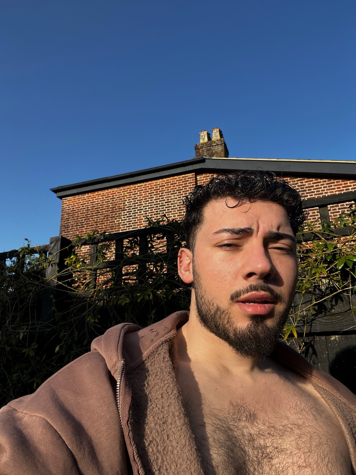 A man poses shirtless in front of a brick building. The photo is taken in natural daylight.