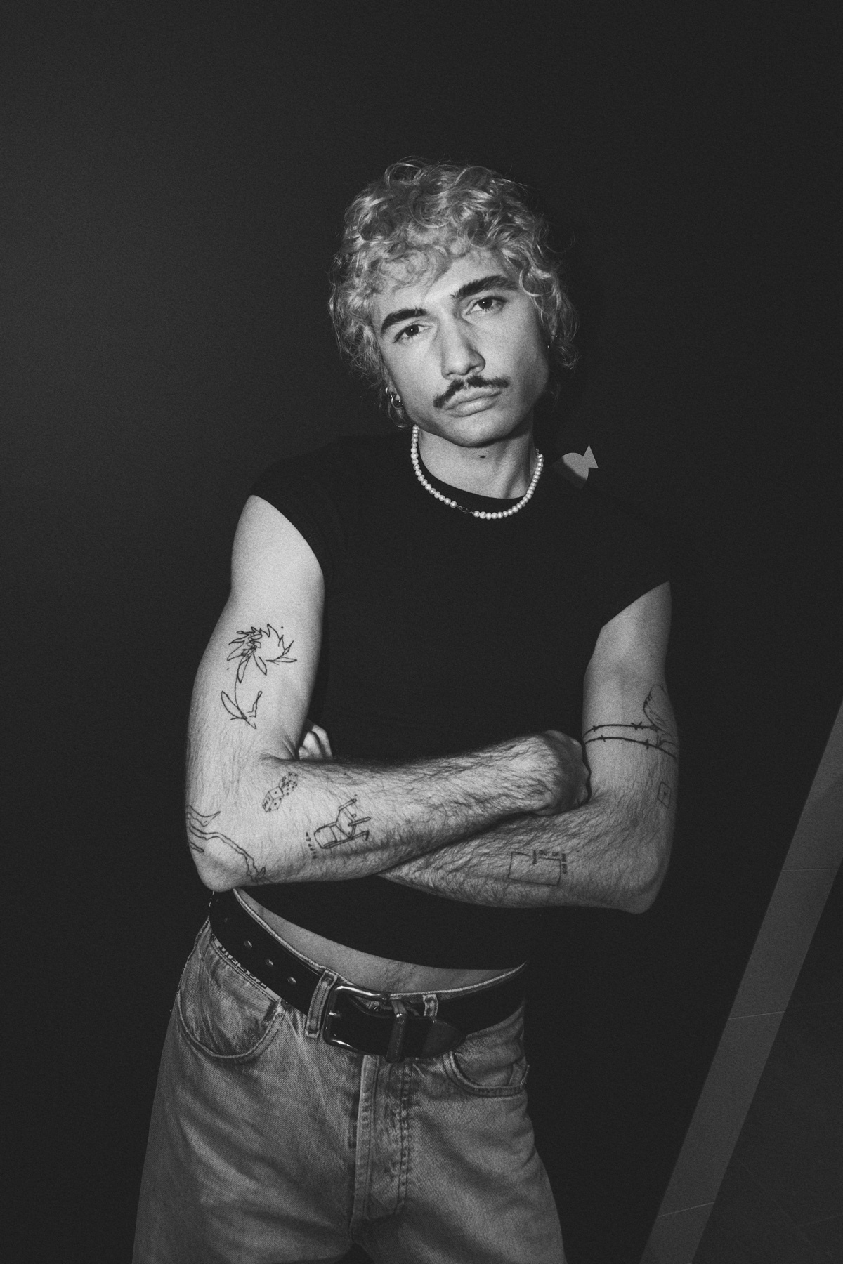 A black and white photo of a man with curly hair and a mustache, wearing a black t-shirt and jeans. He is posing with his arms crossed.