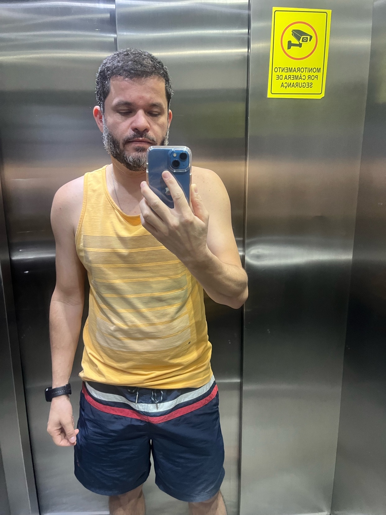 A man takes a selfie in an elevator while wearing a yellow tank top and navy blue shorts. A sign indicating a security camera is visible on the wall behind him.