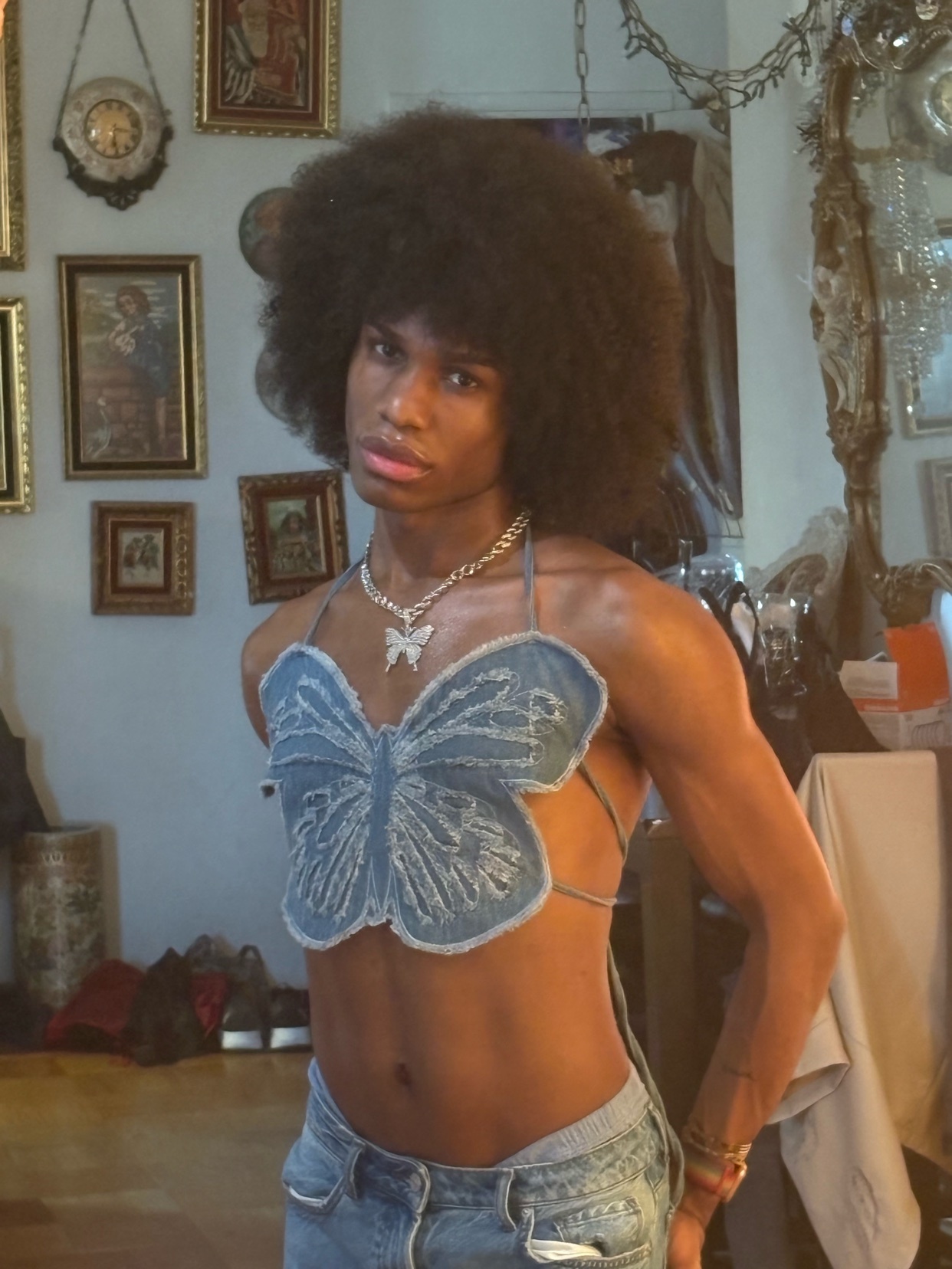A man poses shirtless, wearing a denim butterfly top and jeans. He has an afro and is wearing jewelry.