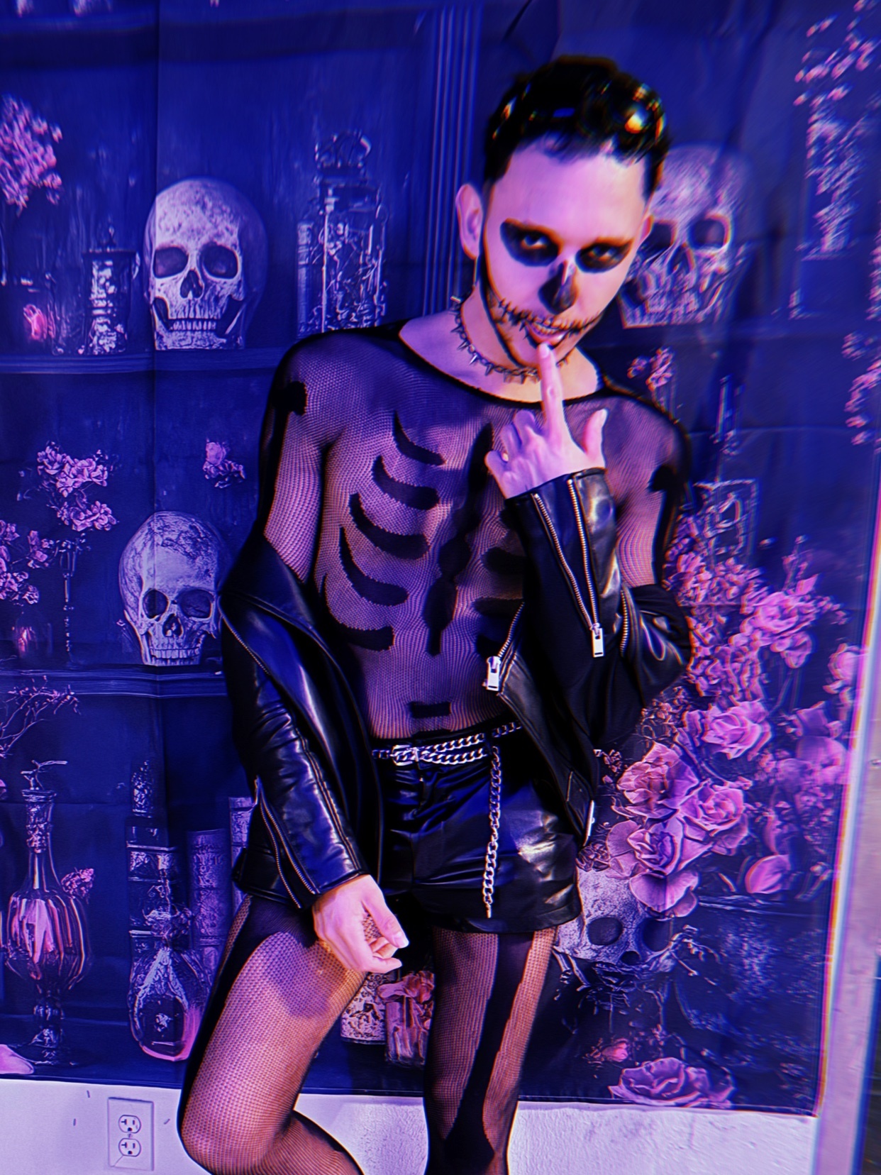 A man in a skeleton costume poses for a photo. He is wearing a sheer top, leather jacket, and shorts.