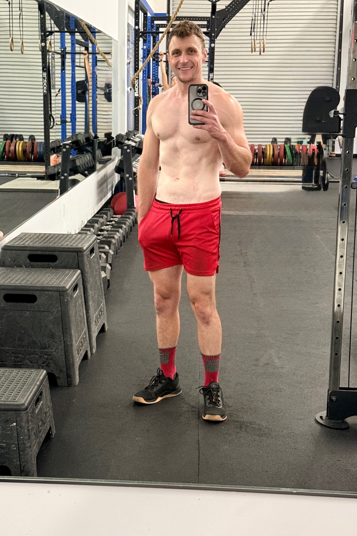 A man takes a mirror selfie in a gym, wearing red shorts and athletic socks. He is shirtless, showing off his muscular physique.