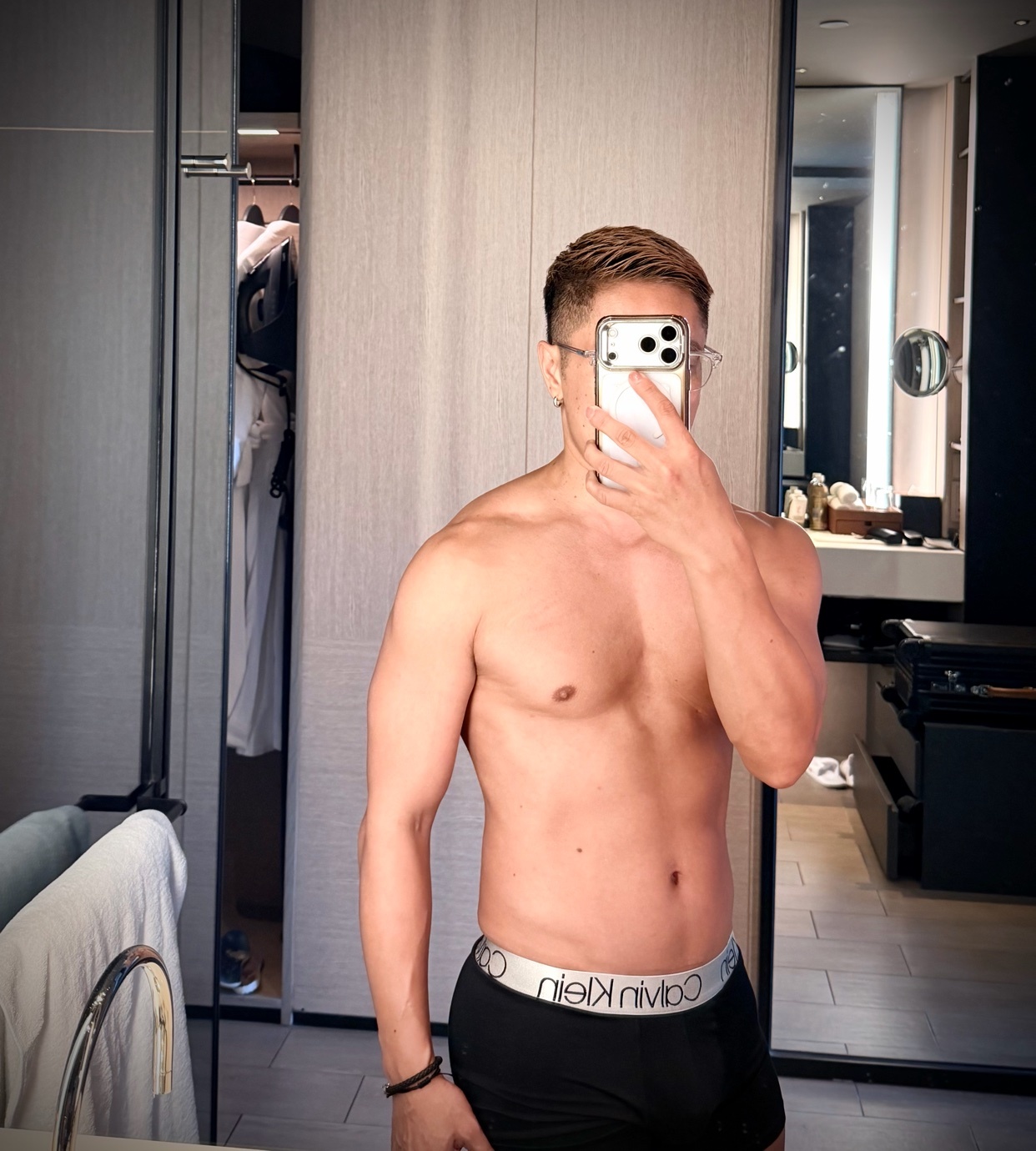 A man takes a mirror selfie in a hotel room, wearing Calvin Klein underwear. He is shirtless and shows off his muscular physique.