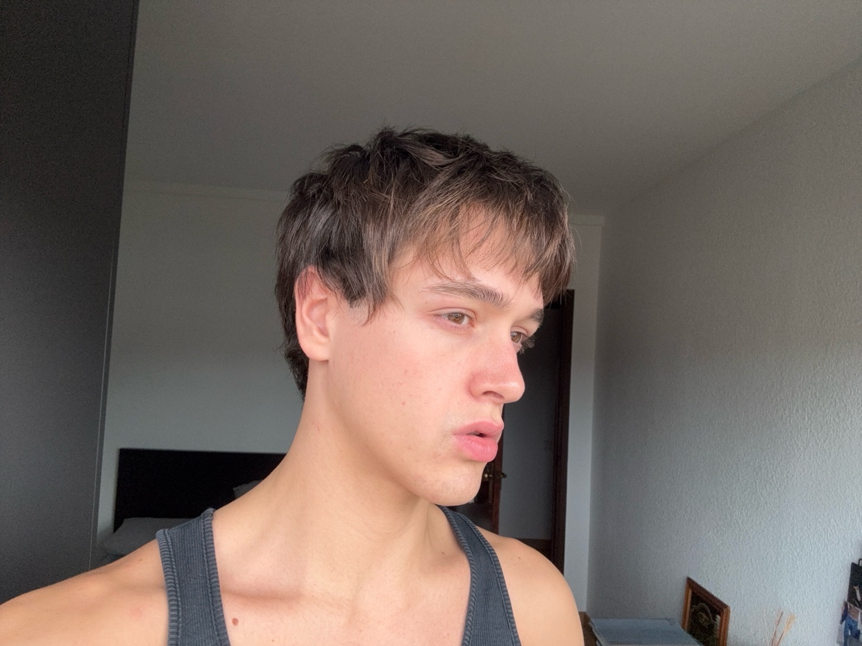 A young man poses for a selfie, looking away from the camera. He is shirtless and wearing a tank top.