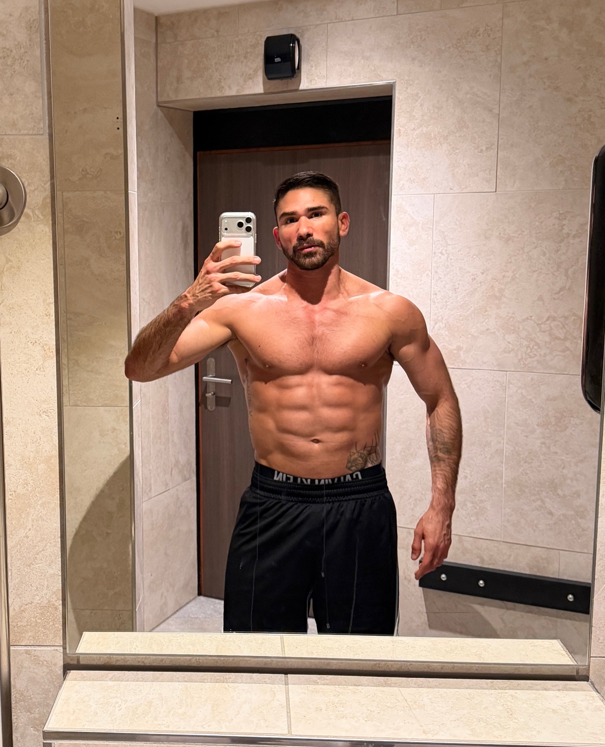 A man takes a mirror selfie, showcasing his muscular physique. He is shirtless, with visible abs and a tattoo.