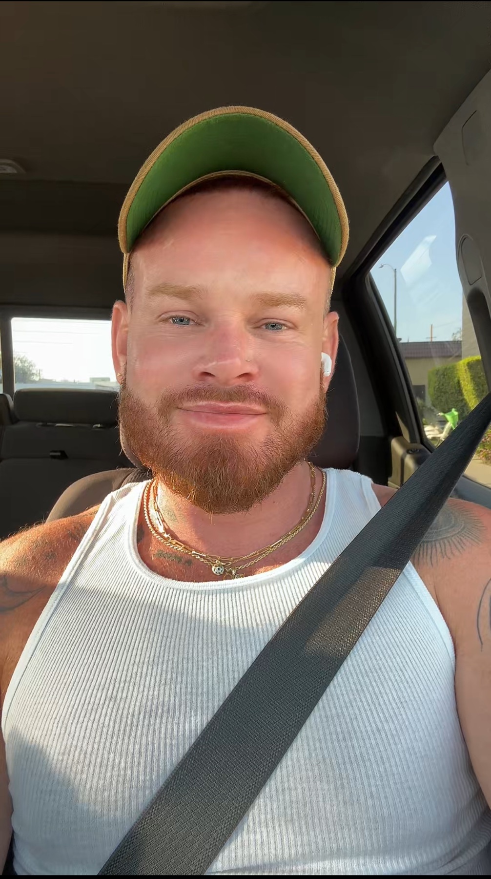 A man with a beard and stubble smiles at the camera while wearing a white tank top and a baseball cap. He is sitting in a car with a seatbelt on and earbuds in.