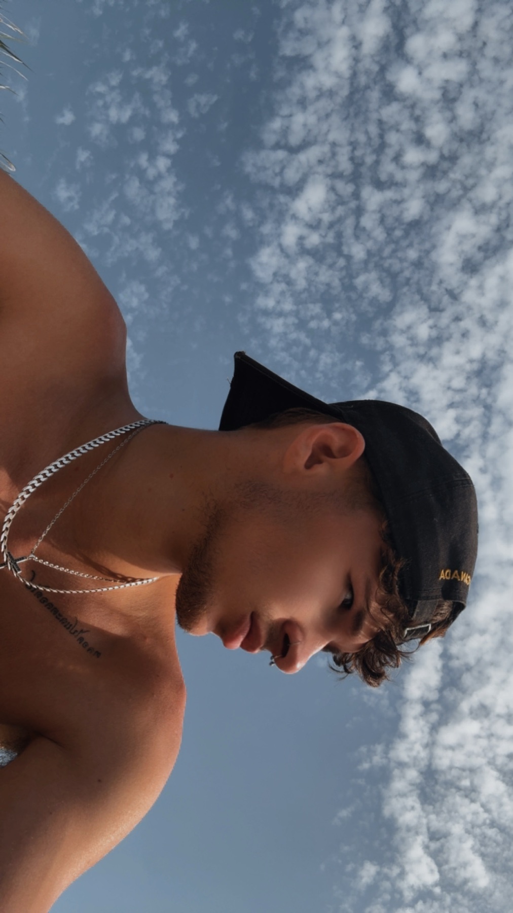 A shirtless man wearing a baseball cap and necklace looks down against a backdrop of a cloudy blue sky. The low-angle shot emphasizes the sky and the man's profile.
