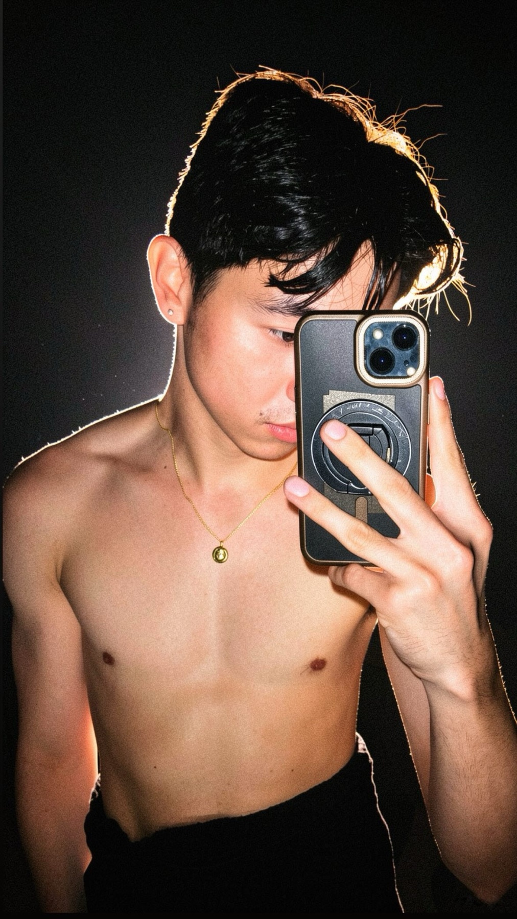 A young man with dark hair takes a selfie in front of a dark background. He is shirtless and wearing a simple gold necklace.
