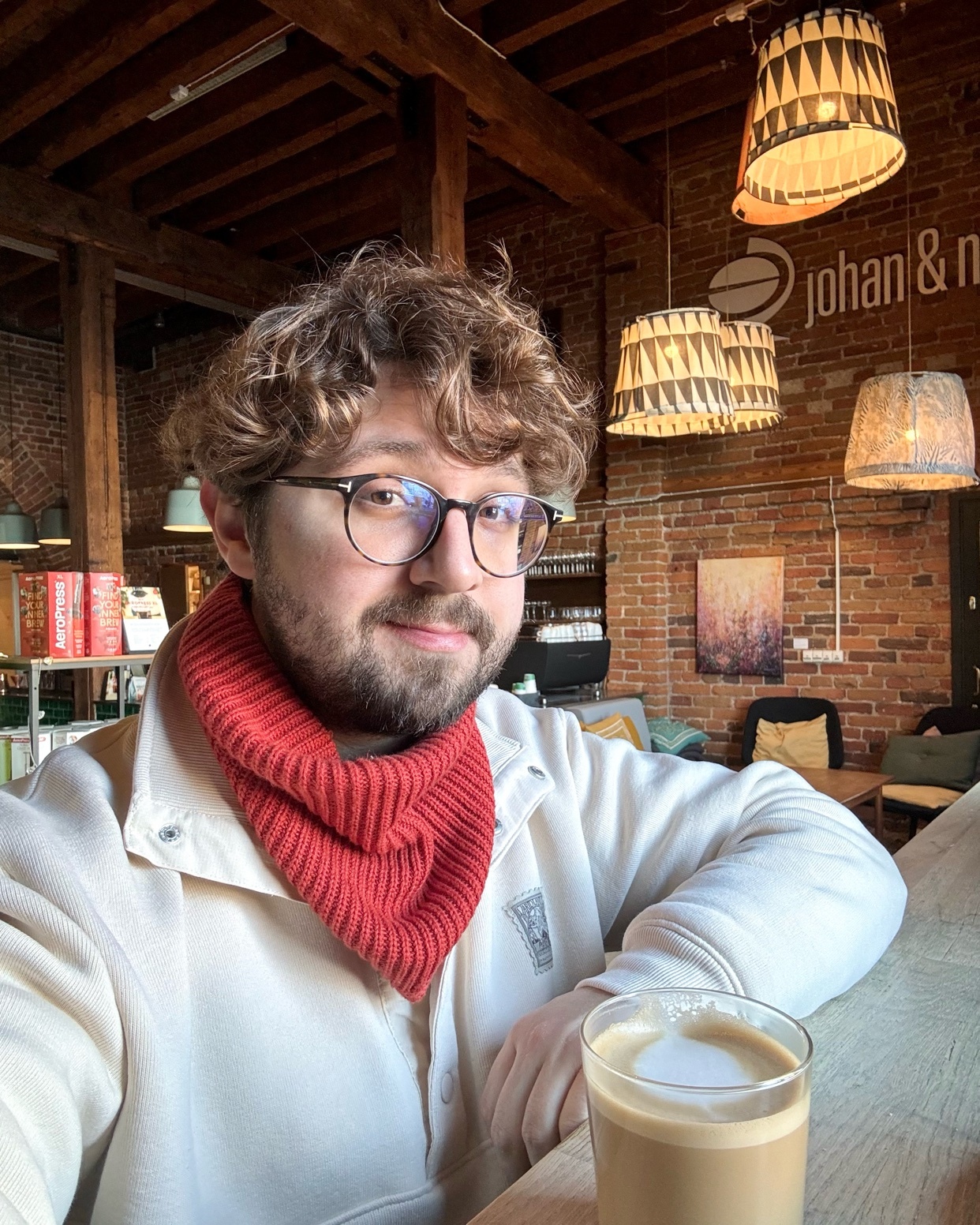 A man with curly hair and glasses smiles at the camera while sitting in a cafe. He is wearing a red scarf and a white jacket.