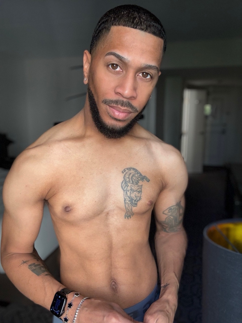 A shirtless man poses for a selfie. He has tattoos and a muscular physique.