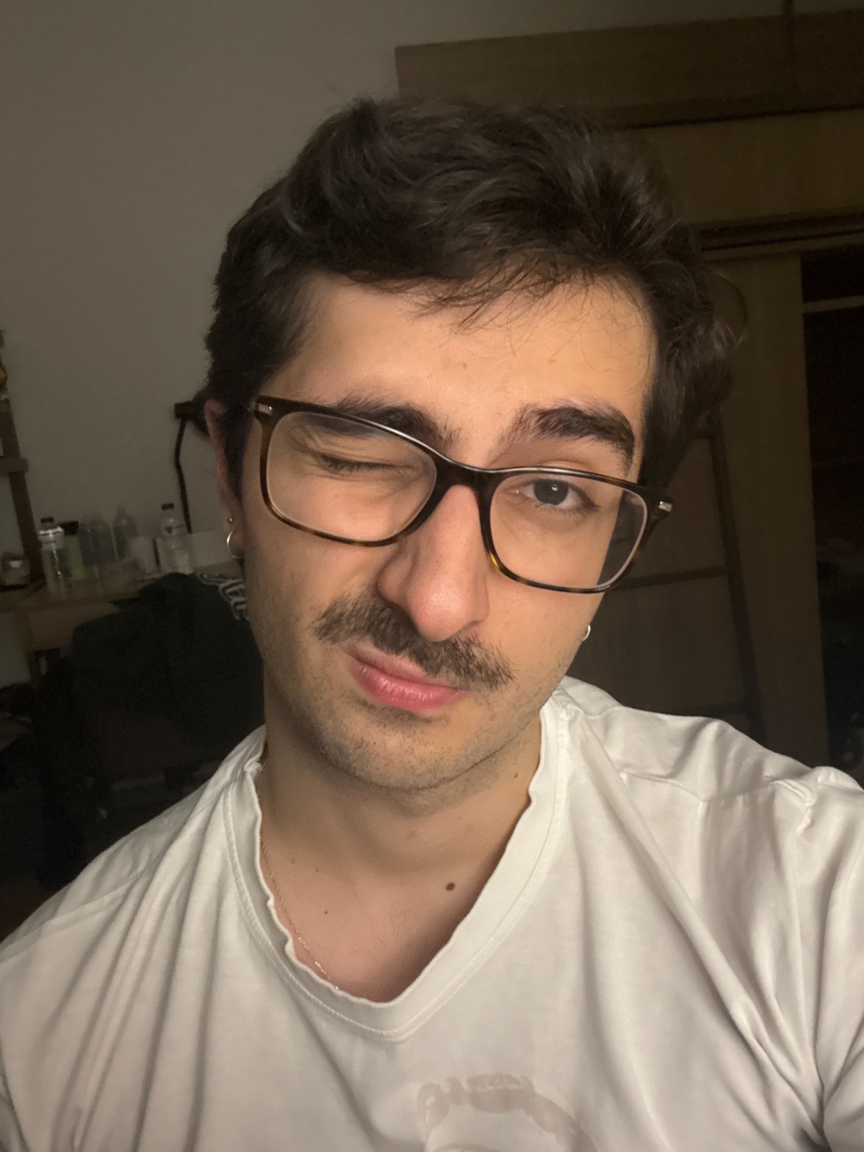 A man winks at the camera, wearing new glasses. He has dark hair and a mustache.