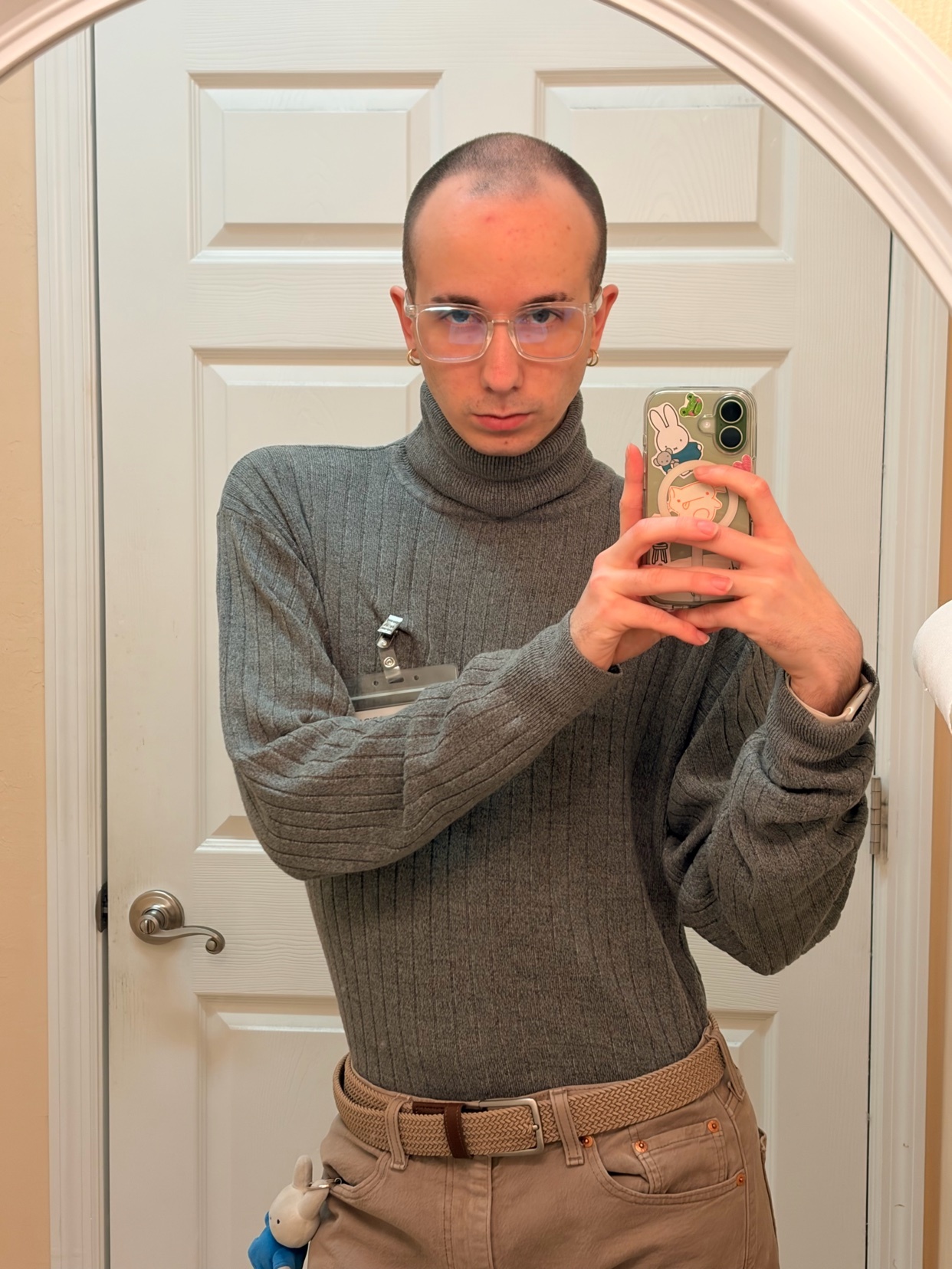 A man in a grey turtleneck and tan pants takes a mirror selfie in a bathroom. He is wearing clear-framed glasses and has short, dark hair.