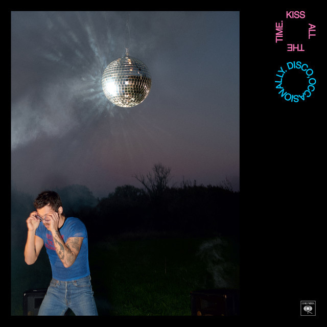 A man with tattoos on his arms and chest wears a blue t-shirt and jeans. He is looking down with his hands near his face, as if shielding his eyes. A disco ball hangs in the air above him, reflecting light. Smoke drifts around the disco ball and in t